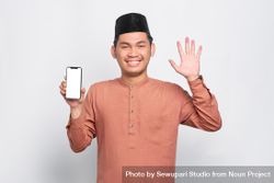 Happy Muslim man in kufi hat with mobile phone waving with other hand 49QZW4