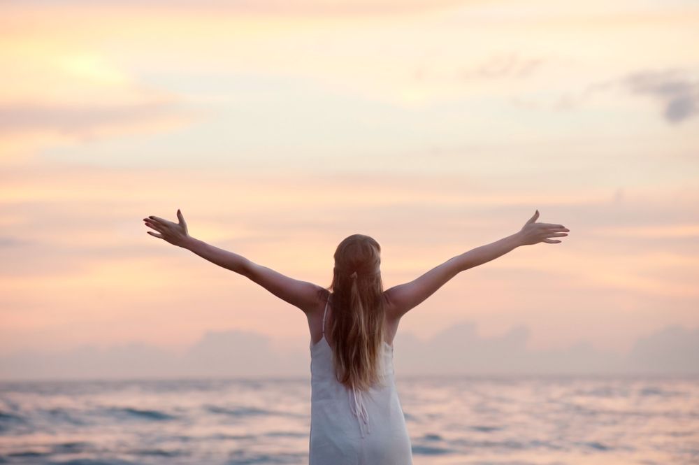 Back view of woman in light dress spreading both arms and standing by seashore at sunset