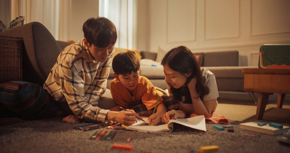 Happy Korean Family At Home: Cute Little Boy Drawing In Notebook On The Floor And Showing His Paintings To Loving And Supportive Mother And Father.