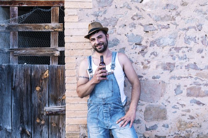 Man in overalls leaning on a wall and holding a beer