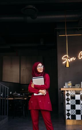 Woman in hijab holding a laptop with both hands and looking away while standing at a cafe