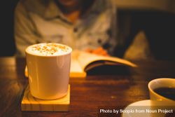 Person sitting at a table with open book and in focus mug of coffee with foam bDWPk4