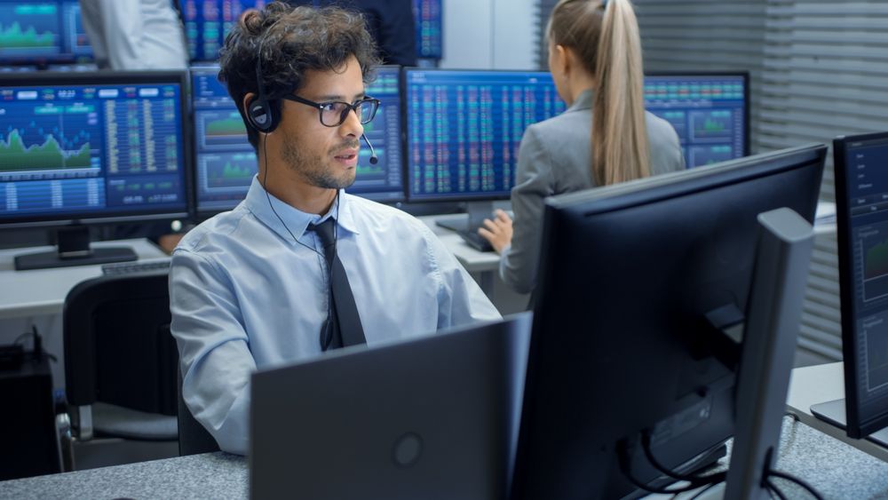 Stock Trader Making Sales with a Headset. In the Background Stock Exchange Office and Group of Traders Working at their Workstations.