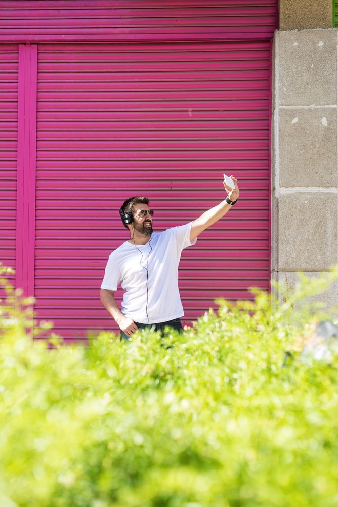 Man in headphones taking selfie with phone in front of pink wall