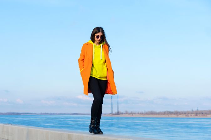 Woman in sunglasses, orange coat, yellow hoodie and jeans against the sea background