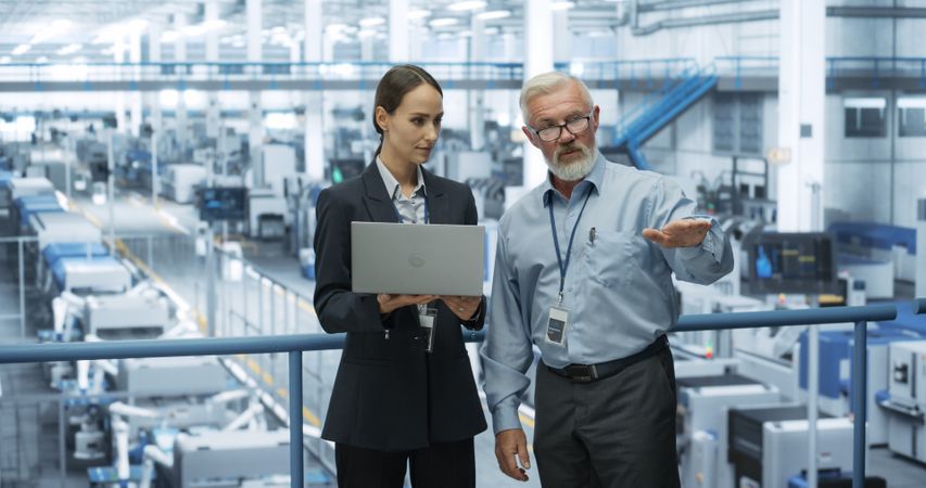Male Engineer and a Female Technician Having a Conversation and Using Laptop Computer. Specialists Monitoring, Analyzing Conditions at a Modern Electronics Factory with Automated Robotic Arms