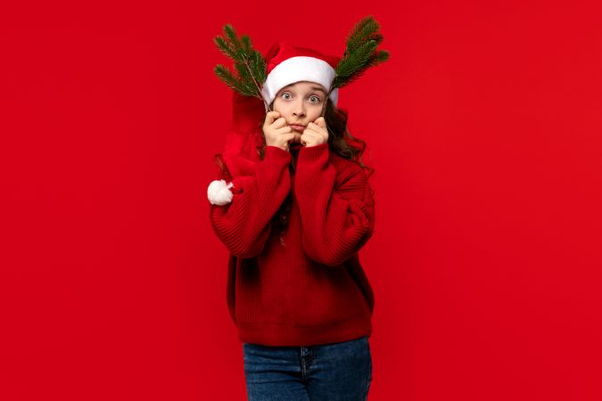 Girl in Santa hat making funny face with fir branches.