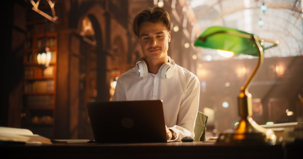 Young Male Student Engaged in Online Education, Using Laptop and Wearing Headphones in a Classical Library