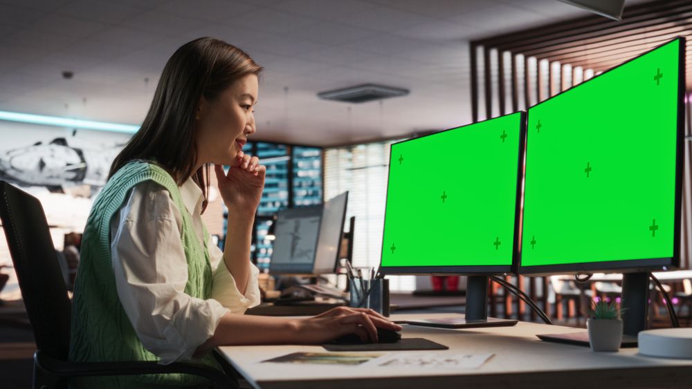 Female Asian Game Designer Using Desktop Computer With Green Screen Chromakey on Display, Designing Characters In 3D Modeling Software For Survival Video Game.