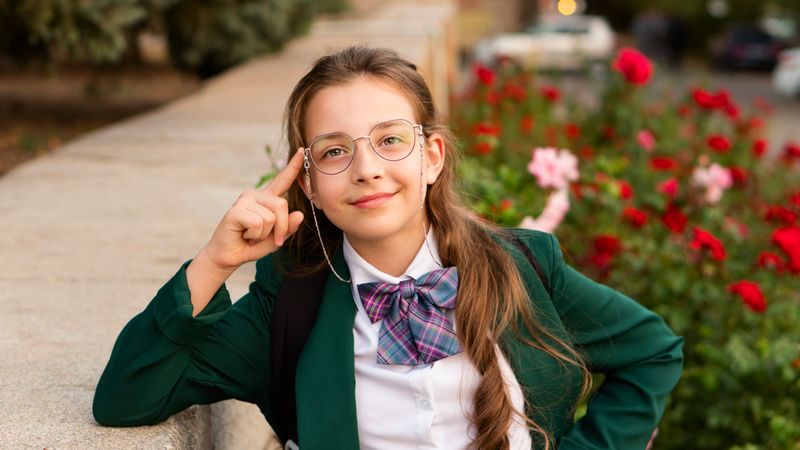 Girl in uniform smiling thoughtfully with hand raised by glasses.