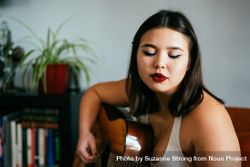 Horizontal shot of young woman sitting inside home playing guitar 4Zedx5