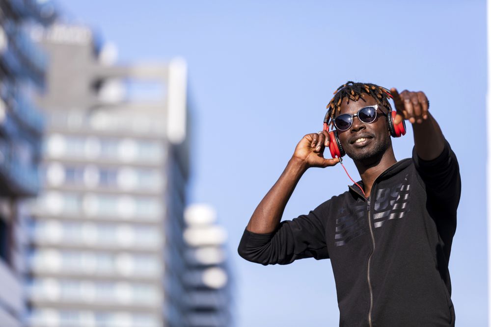 Smiling Black man wearing headphones & sunglasses standing on the street listening to music while pointing ahead