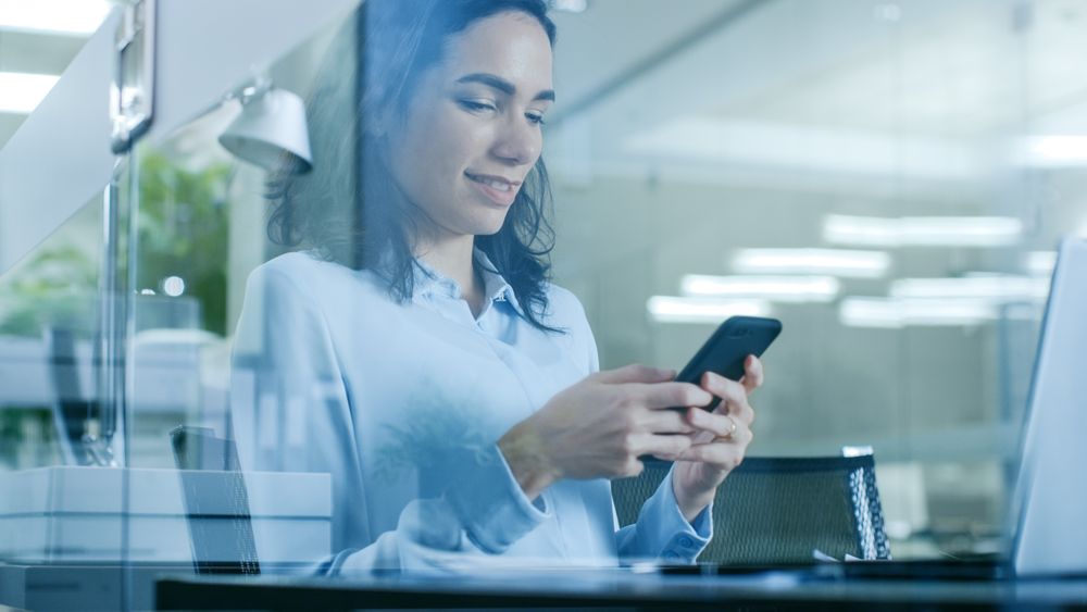 Female Office Worker Uses Smartphone while Sitting at Her Working Desk with Laptop on it.