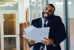Laughing businessman in suit and tie with papers walking in office while speaking on smart phone