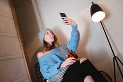 Woman in sweater and hat taking selfie and sitting near wall