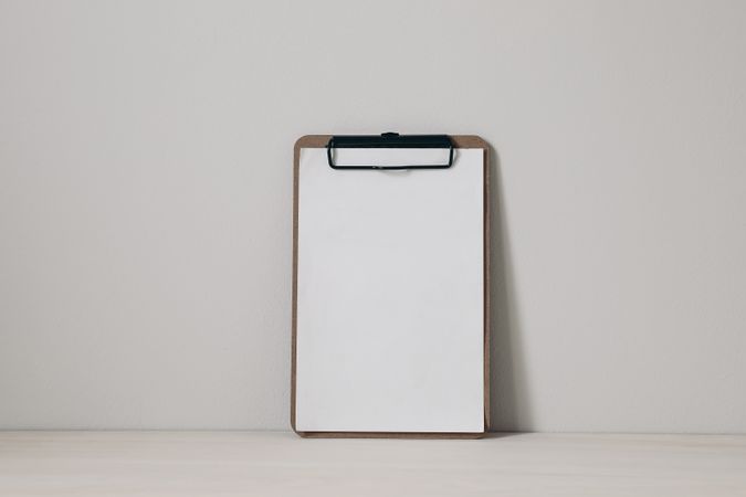 Wooden clipboard mockup with blank paper sheet on a wooden table against neutral beige wall background