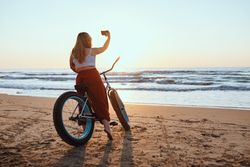 Cheerful woman taking selfie on smartphone while resting with bicycle at sunny shore