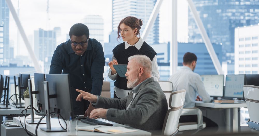 Diverse Colleagues Talking In Front Of Desktop Computer In Corporate Office With Megapolis Window View.