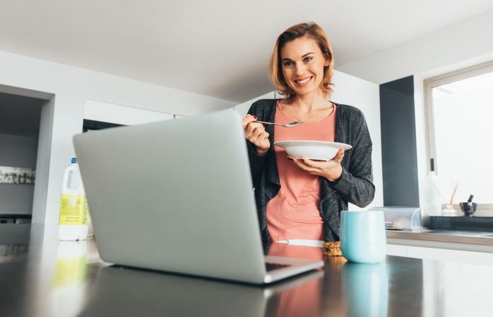 Woman smiling at laptop with cereal bowl in kitchen