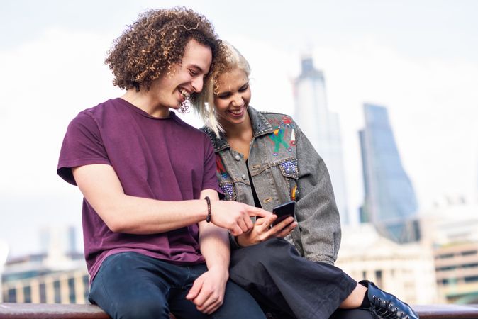Smiling blonde woman and curly haired man looking at phone while sitting on fence over river