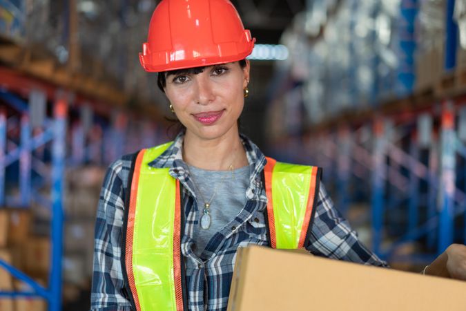 Woman in hard hat carrying in box in warehouse