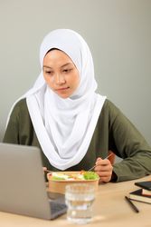 Happy Asian Muslim Woman is eating Her Healthy Salad while Working on Her Laptop.