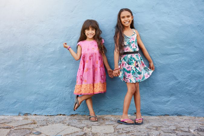 Full length portrait of two happy little girls standing together against blue wall