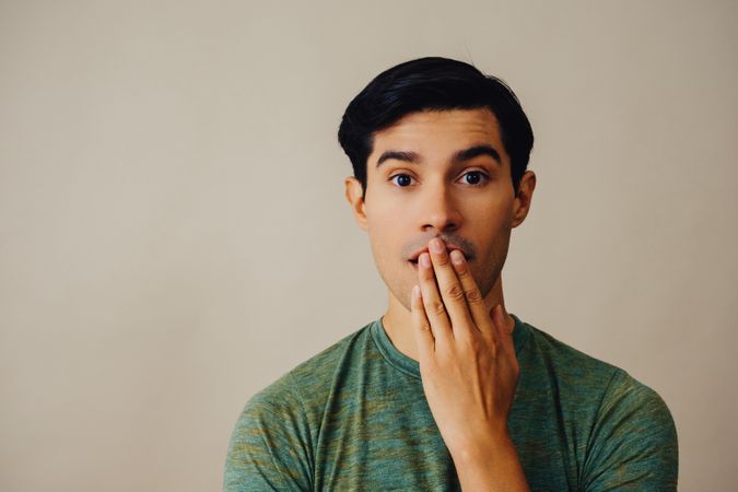 Portrait of Hispanic male in neutral room with hand over his mouth, copy space