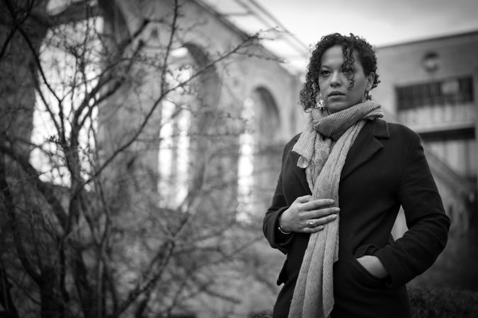 Grayscale photo of woman in coat standing near bare trees outdoor
