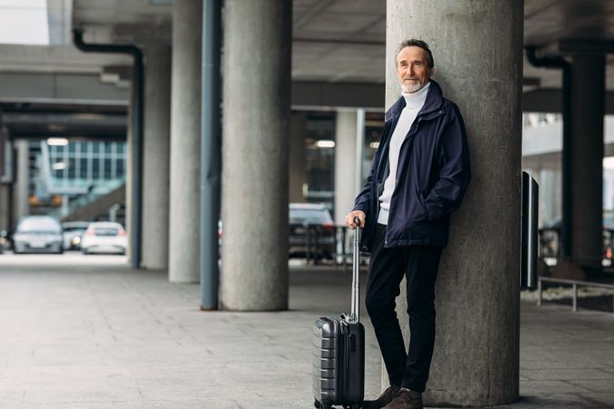 Mature man with suitcase leaning against concrete pillar