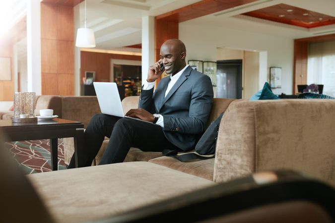 Male executive waiting in hotel lobby using cell phone and working on laptop