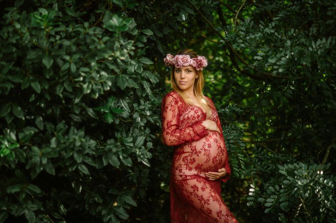 Pregnant woman with closed eyes touching belly and chin while standing in garden in sunny day