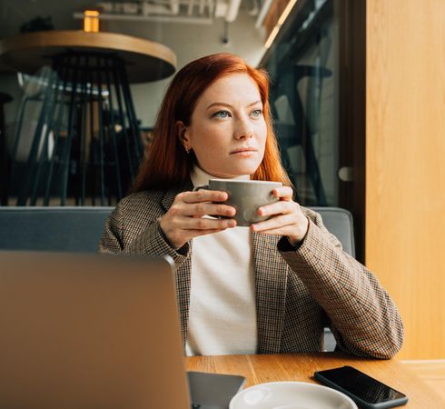 Red haired woman enjoying a coffee while sitting at laptop in cafe
