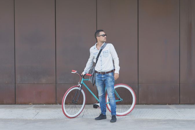 Male in sunglasses standing with red and green bicycle parked against a brown wall, vertical