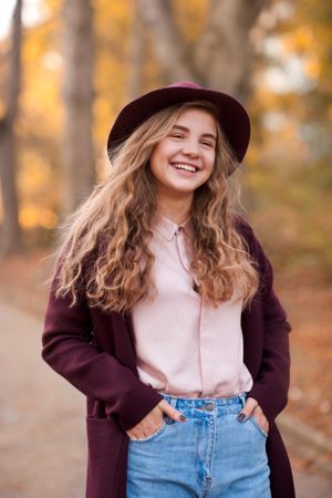 Cheerful teenage girl in purple coat with hat standing near fallen autumn tree leaves