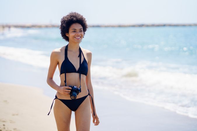 Confident smiling woman walking along the shore in bikini with camera around her neck