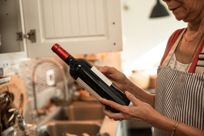 Cropped image of older woman holding a bottle of red wine in kitchen