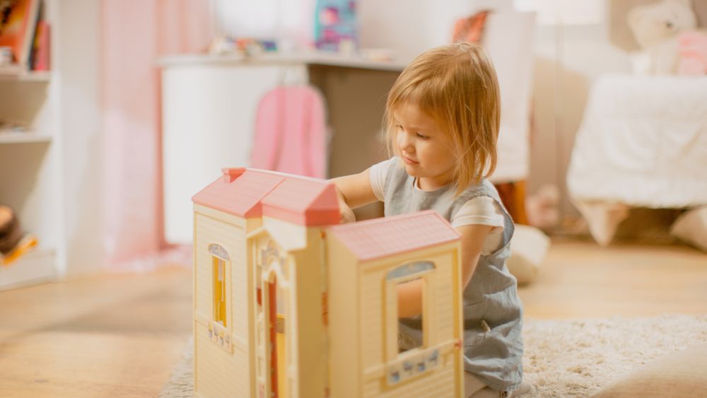 In the Children's Room: Happy Little Girl Playing with Doll House and Sitting on a Carpet.
