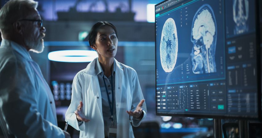 Neuroscience Laboratory Meeting: Female Asian Scientist And Male White Surgeon Discussing MRI Brain Scan On TV Display.