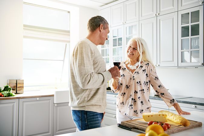 Grey haired couple in kitchen toasting with wine over a breadboard with cheese