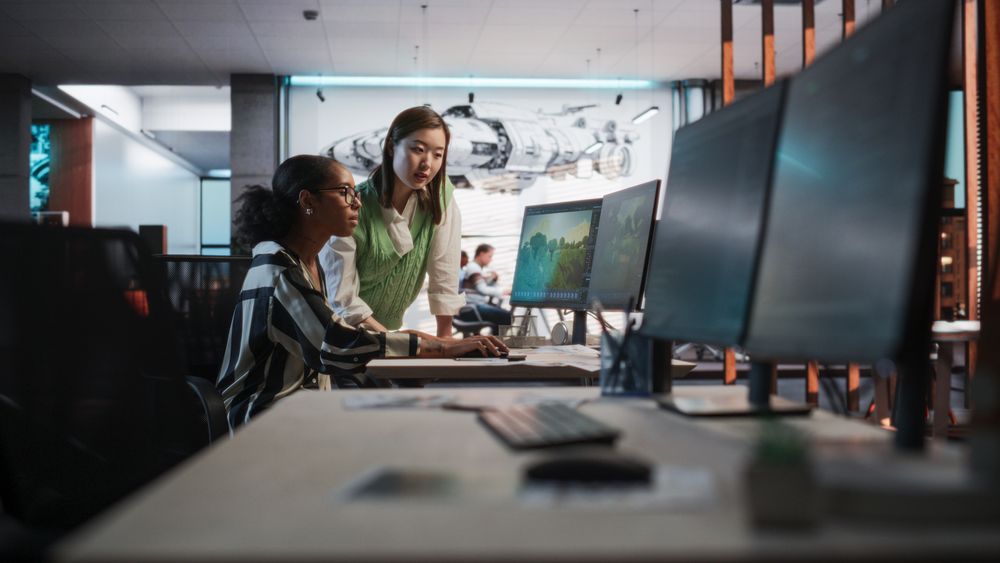 Black Female Game Developer Using Desktop Computer With 3D modelling Software And Talking to Asian Female Project Manager in Game Design Studio Office.