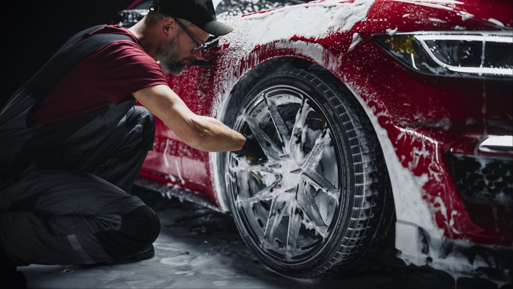 Car Ad Style Photo of a Professional Car Wash Specialist Using a Big Soft Sponge to Wash the Rims of a Red Sportscar with Shampoo Before Detailing,