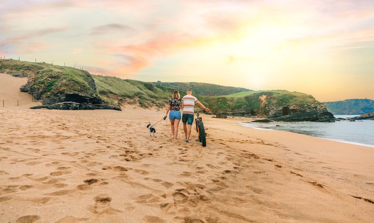 Back of couple strolling with bicycle and small dog along the coast