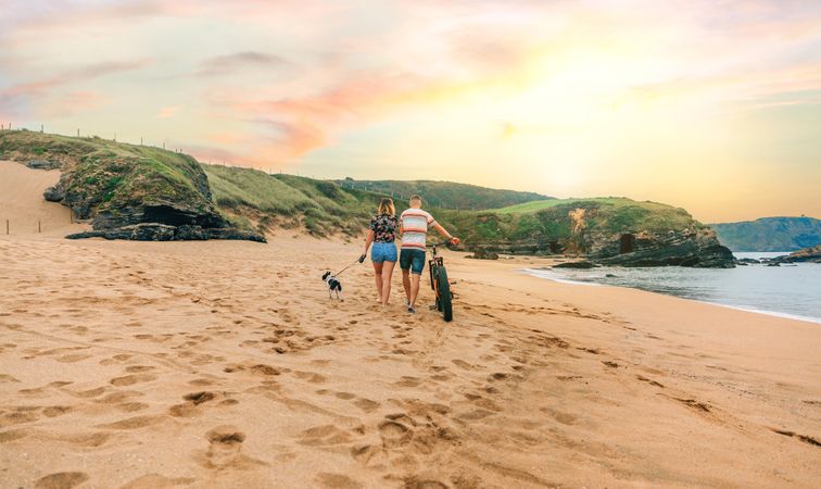 Back of couple strolling with bicycle and small dog along the coast