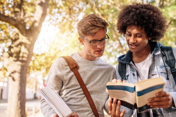 Two college friends reading book together outdoors on school campus