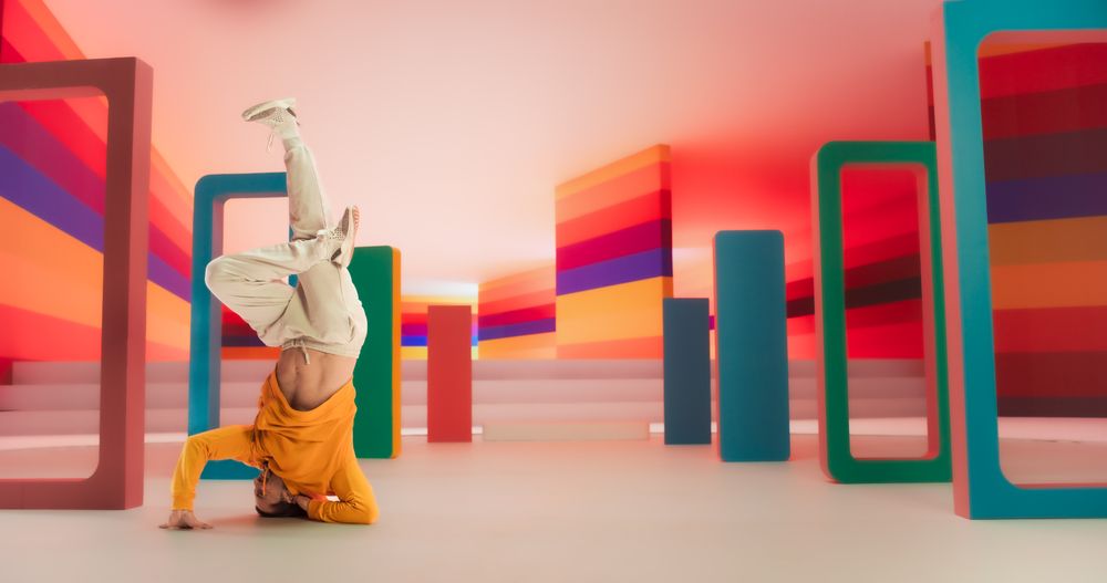 Male Dancer in Bright Sweater Performing Swift Breakdancing Moves With A Freeze in a Colorfully Decorated Studio. Professional B-Boy Showcasing Talent and Skills, Surrounded by Geometric Decoration