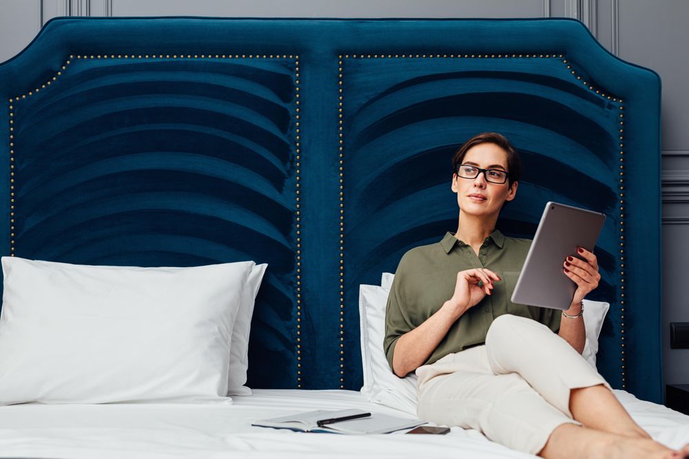 Woman sitting on a bed with a blue headboard working on a tablet