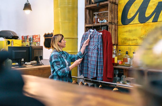 Woman holding digital tablet on hand while checking price label of plaid shirt on clothing shop