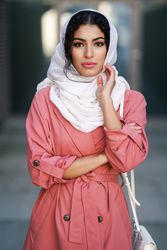 Female in headscarf and pink trench coat standing confidently with with one hand to her face