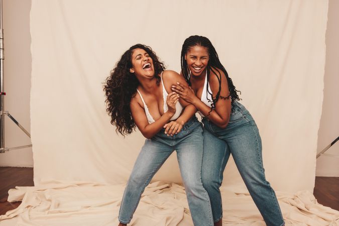 Two cheerful women laughing and being playful while wearing denim jeans in a studio